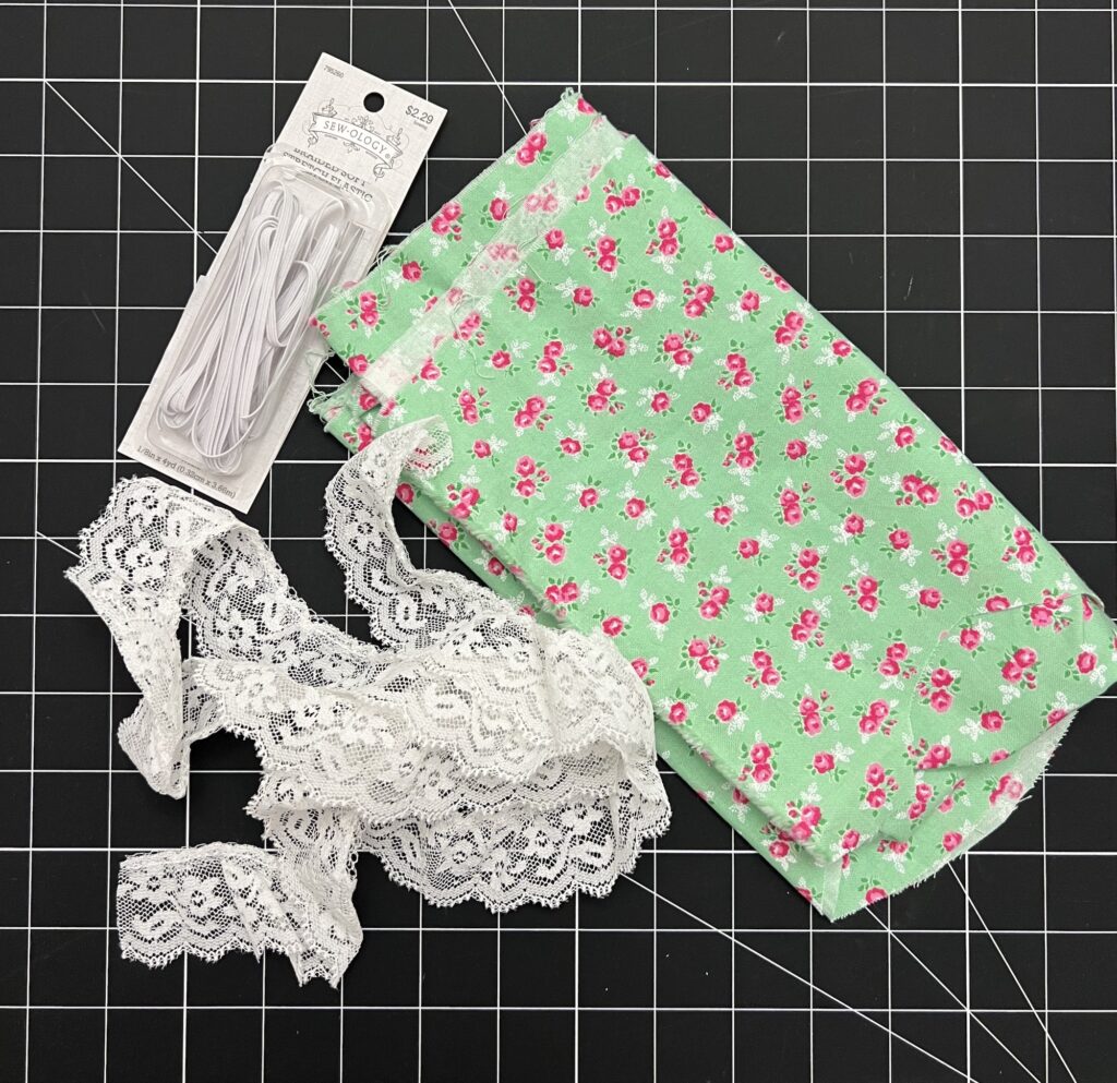 A packet of white elastic, green floral fabric, and white lace rests on a black grid cutting mat, perfect for crafting a tiered skirt with layered elegance.