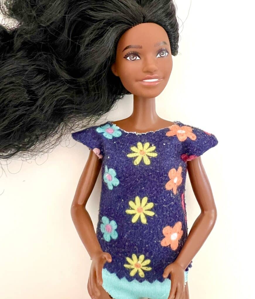 A doll with long black hair is wearing a colorful flower-patterned dress that complements her stylish Barbie shirt, standing gracefully against a white background.