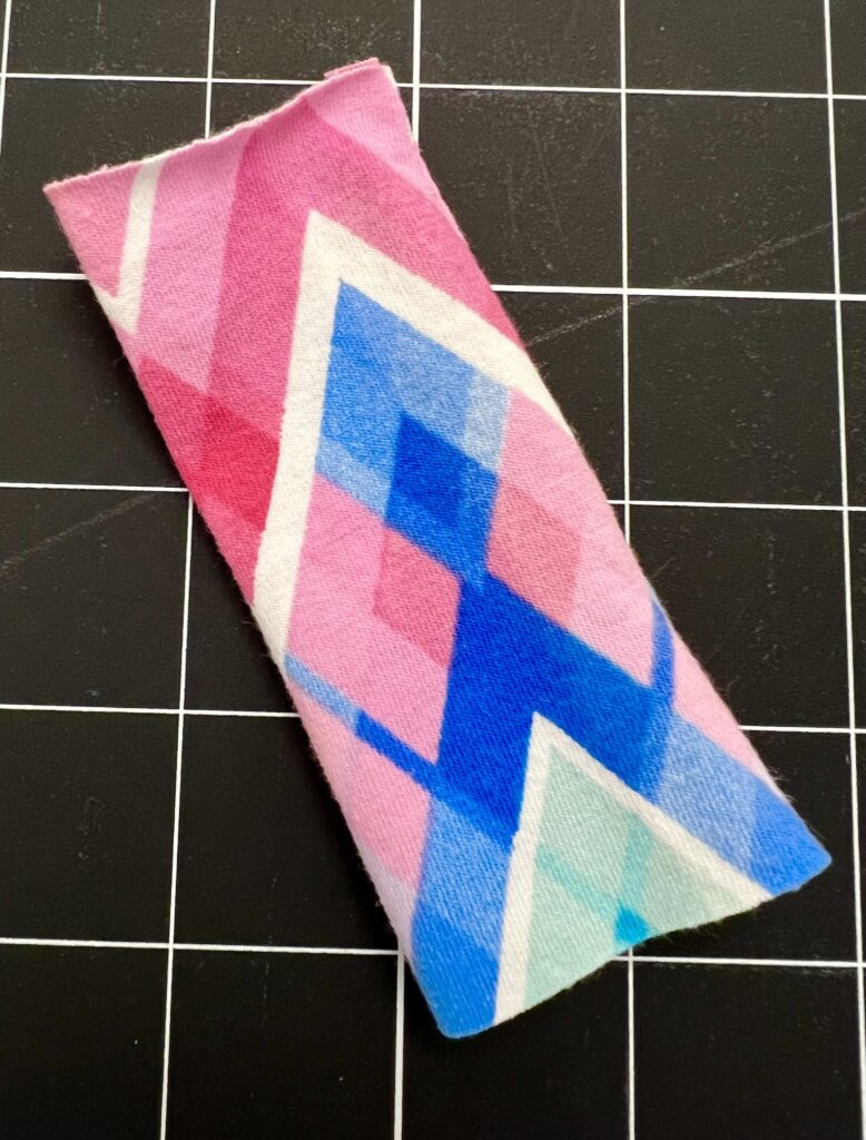 A rectangular fabric piece with a zigzag pattern in pink, blue, and white on a black and white grid background.