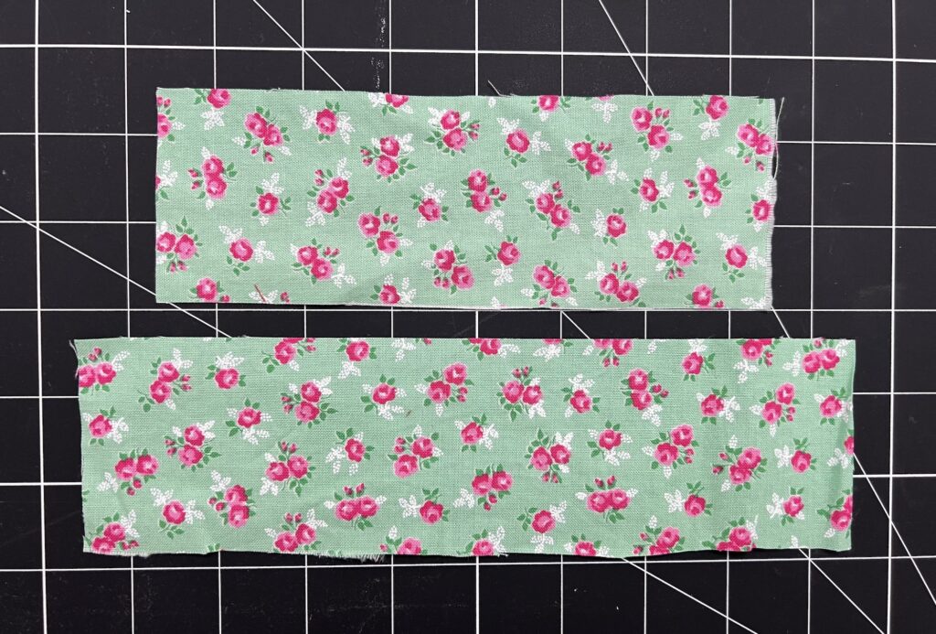 Two rectangular pieces of green fabric with a pink floral pattern lay gracefully on a black cutting mat grid, ready to be transformed into a charming tiered skirt.