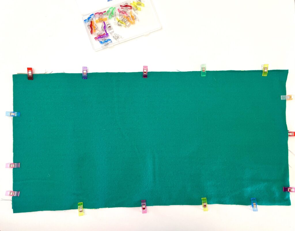 A rectangular piece of green fabric is held in place by multicolored clips around its edges. A small box of additional clips is visible in the top left corner.