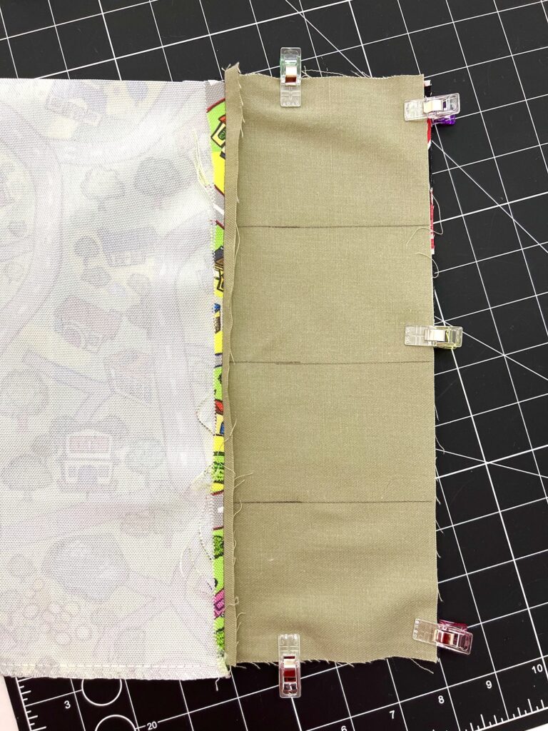 Two pieces of fabric, one with a colorful pattern and one solid beige, are clipped together with sewing clips on a black cutting mat.