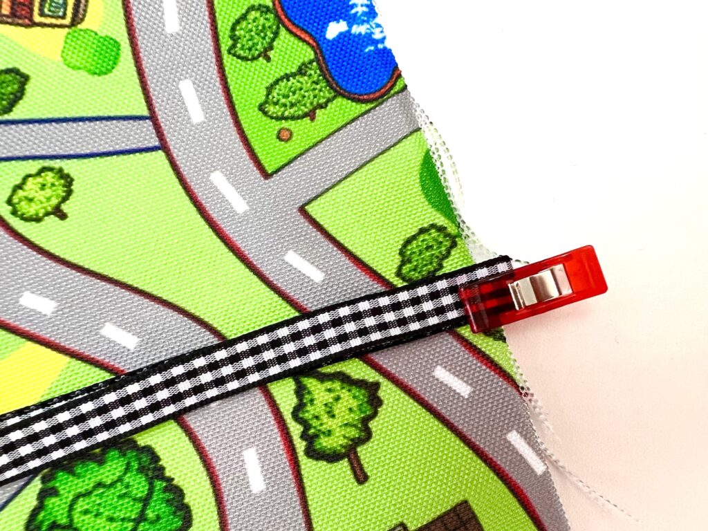 A close-up of a colorful fabric depicting a road map with trees and a pond, held together by a red clip and a black-and-white checkered ribbon.