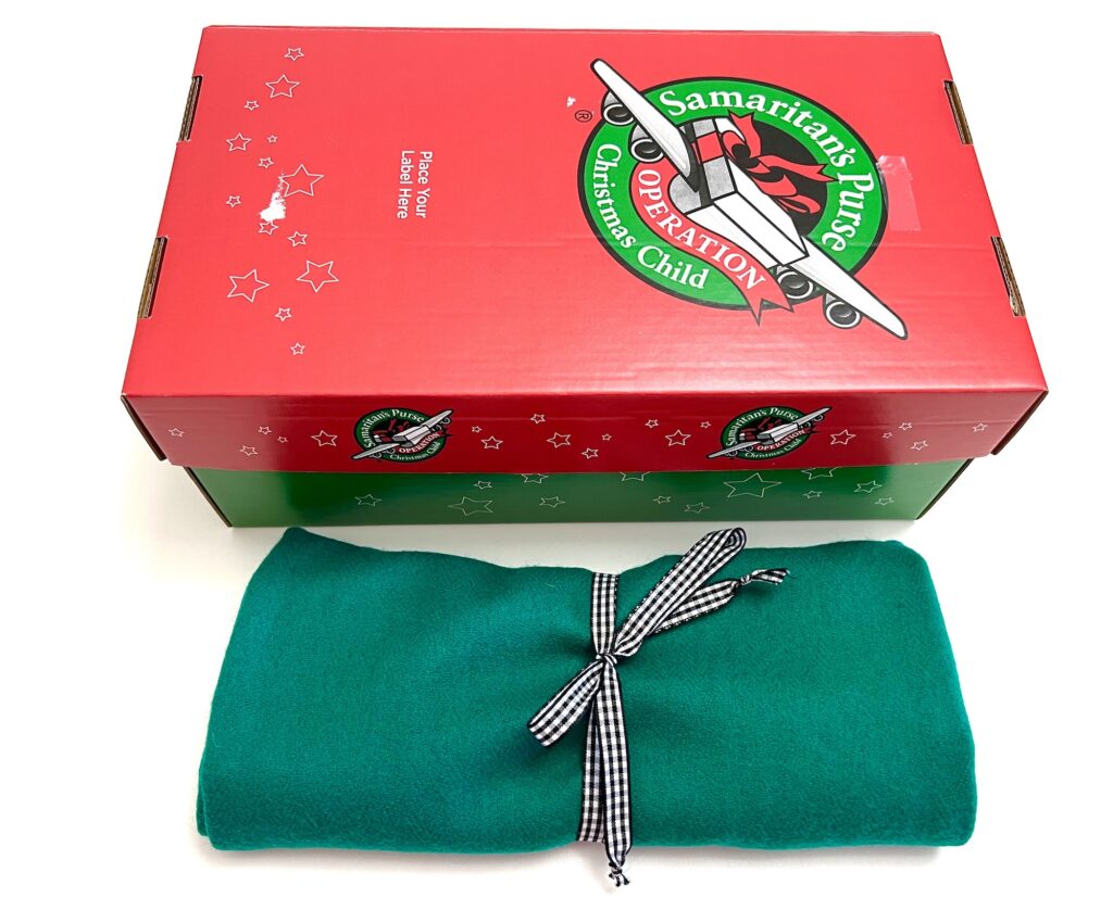 Red and green Samaritan's Purse Operation Christmas Child shoebox with a folded green playmat tied with a checkered ribbon in front.