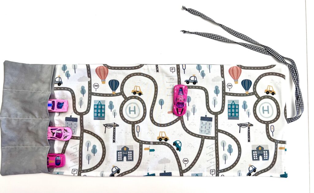 A playmat designed as a fabric road map, complete with small toy cars cruising the pathways. Featuring a built-in gray pouch and a handy drawstring, it's perfect for easy storage after playtime adventures.