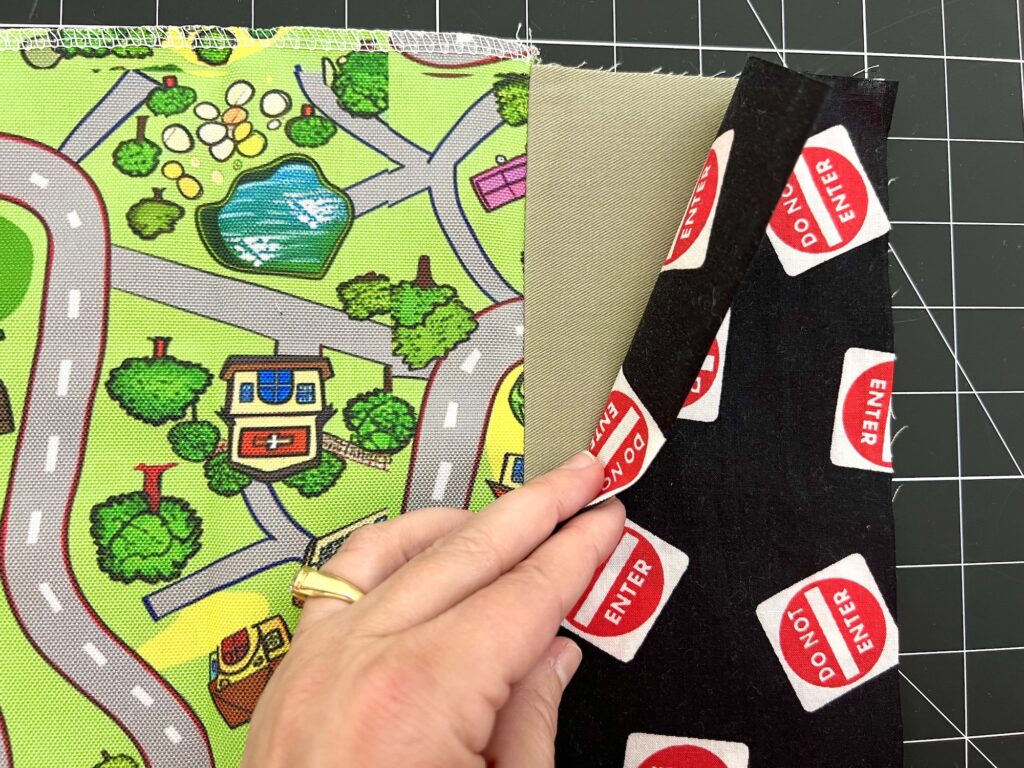 A hand holds two fabric samples. One features a colorful town map design; the other has red "DO NOT ENTER" signs on a black background. A cutting mat is visible underneath.