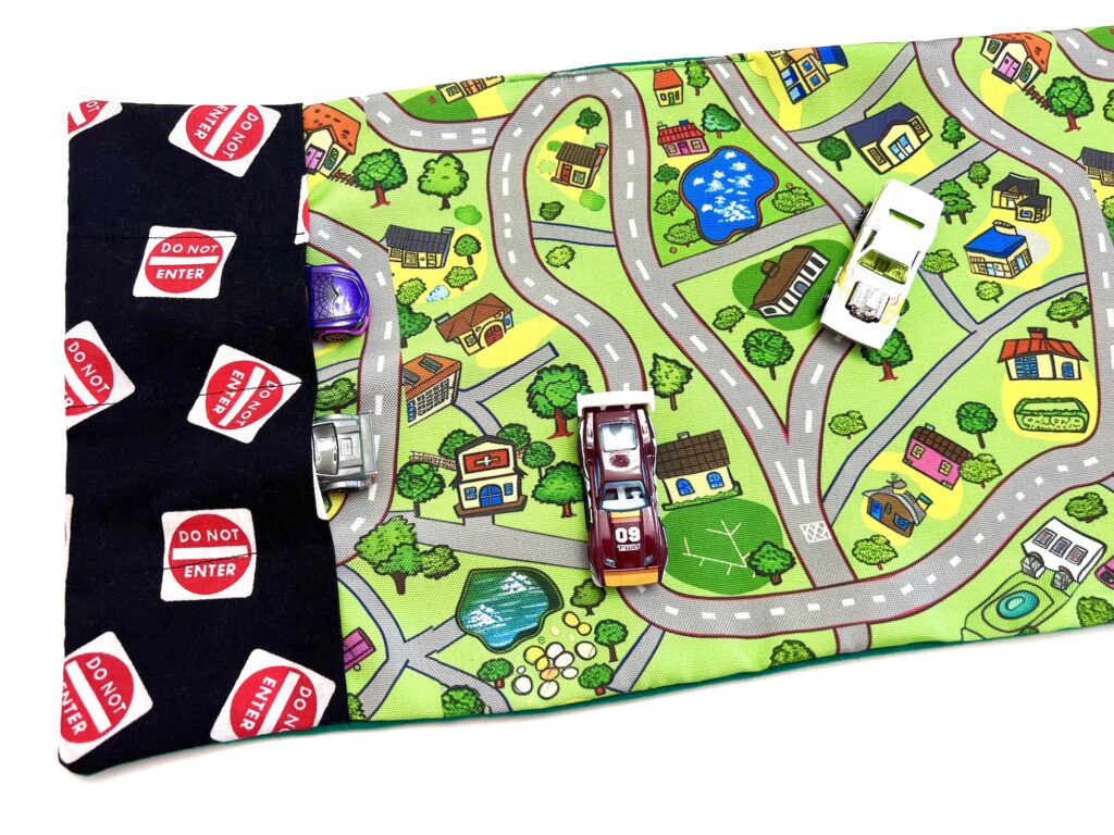 A vibrant playmat showcases toy cars navigating roads, buildings, and greenery. On the left, a black section marked with red "Do Not Enter" signs adds an exciting element to the scene.