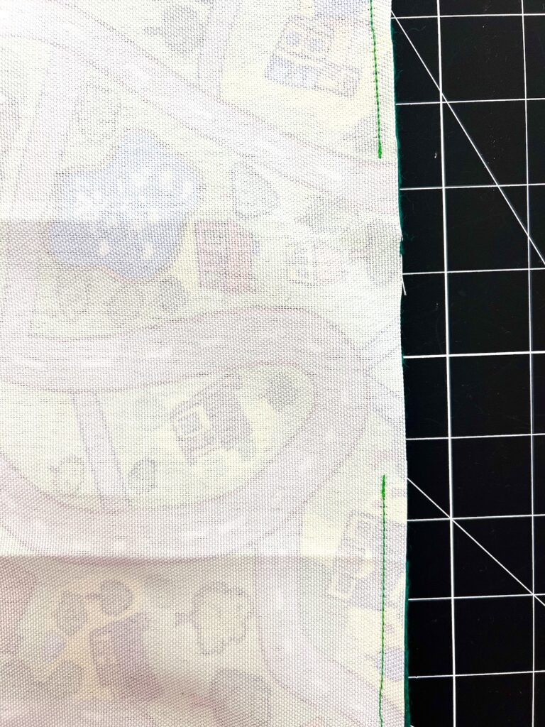 Fabric with a faint road and building pattern is placed on a grid cutting mat, featuring a single green stitched edge on the right.