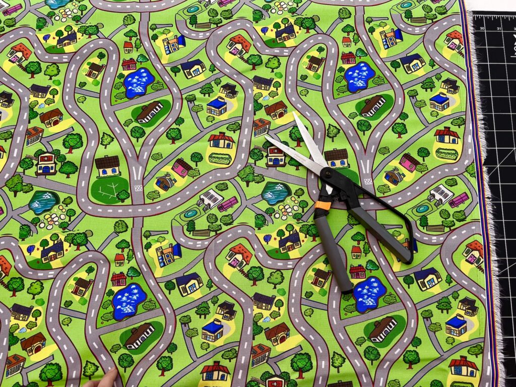 Colorful fabric with a cartoon road map design, featuring winding roads, trees, and buildings. A pair of scissors rests on the fabric, positioned on a cutting mat.