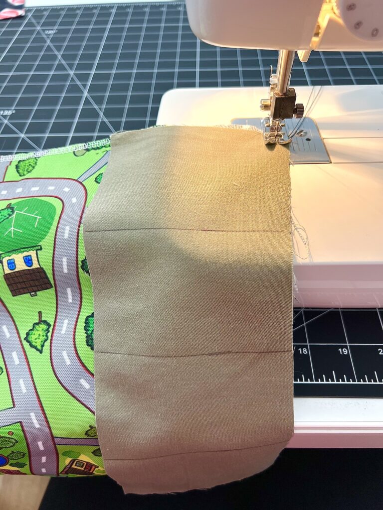 A sewing machine stitches together two pieces of fabric, one beige and the other with a colorful road pattern, on a cutting mat.