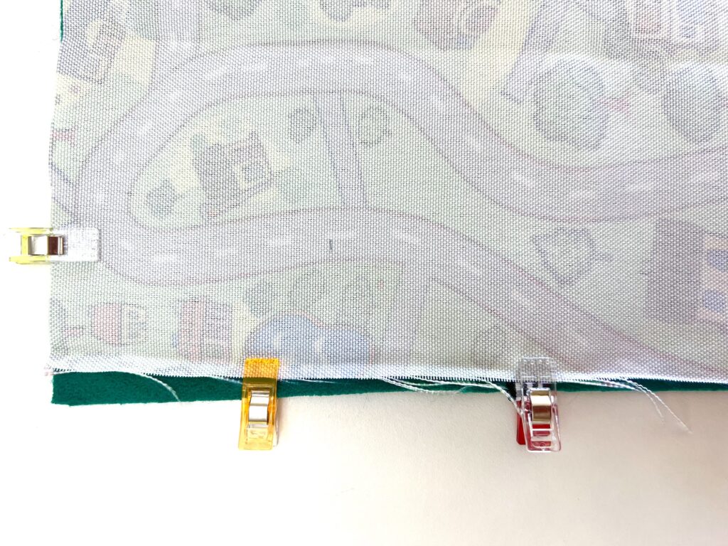 Fabric with a map design is clipped with three sewing clips in yellow, red, and clear colors, on a green surface.