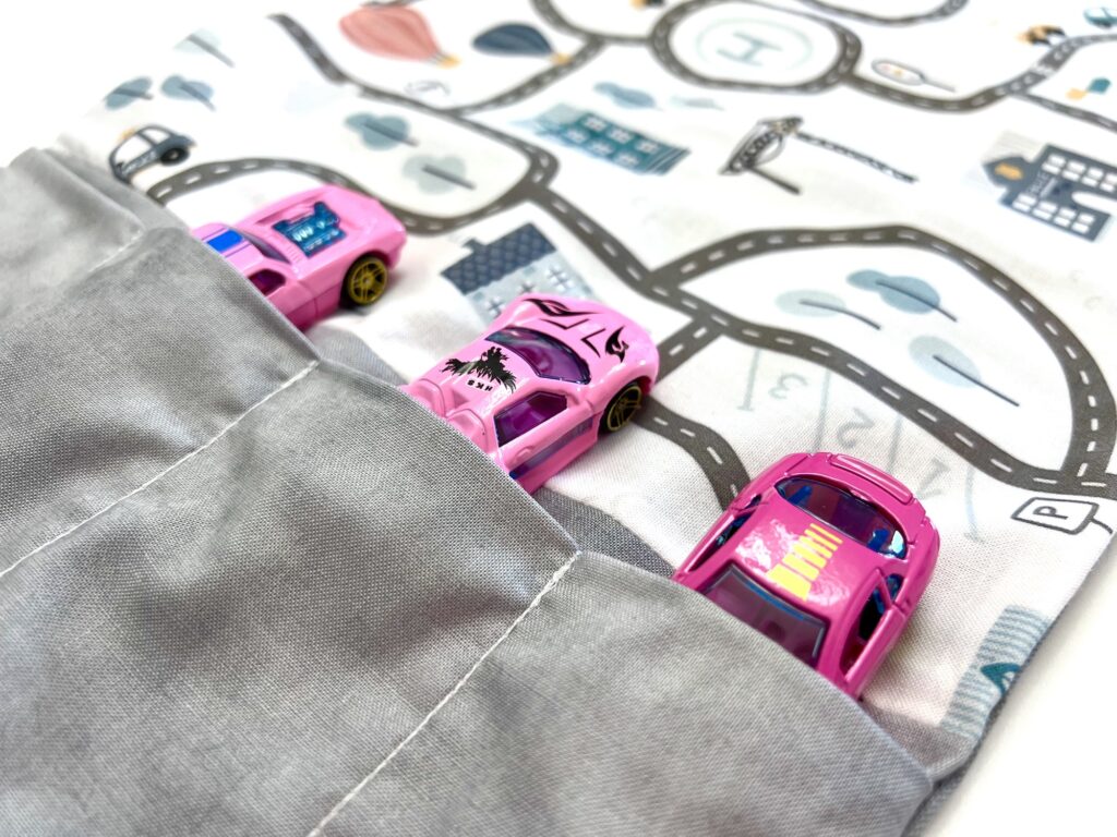 Three pink toy cars are placed in pockets on gray fabric. Above them, a patterned playmat features a road map design with buildings and roads.