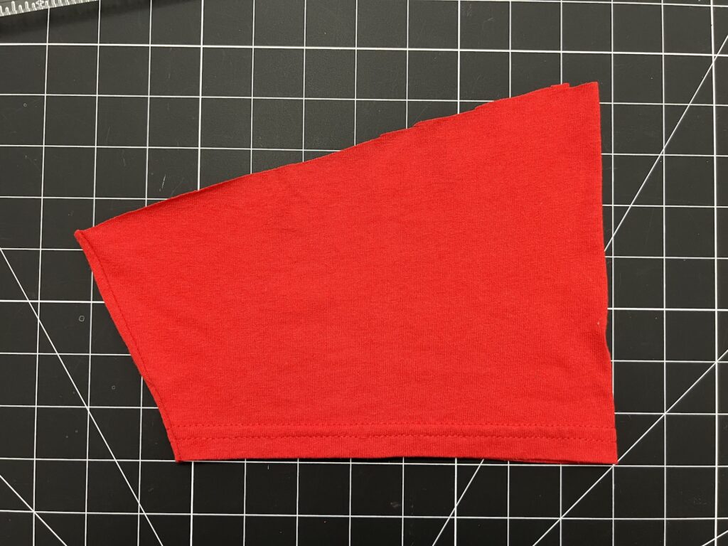 A piece of red fabric, possibly from a t-shirt sleeve, is cut into an angled shape and placed on a black grid cutting mat—perfect for making drawstring bags.