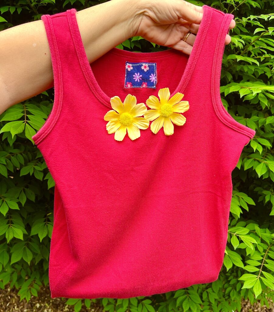 A hand holds a bright pink sleeveless ribbed tank top bag with two yellow artificial flowers attached near the neckline, set against a leafy green background.