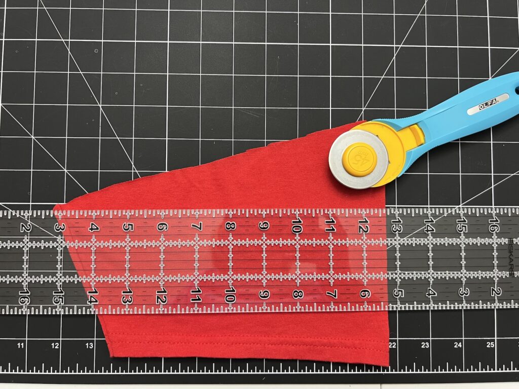 A rotary cutter and clear ruler rest atop a red fabric piece—possibly for t-shirt sleeve drawstring bags—all arranged neatly on a black cutting mat with a white grid.