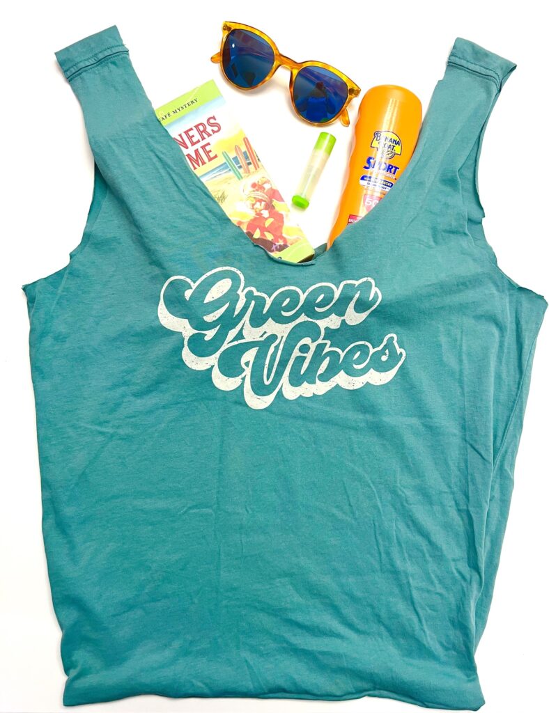A blue tank top with "Green Vibes" text, sunglasses, a book, a t-shirt bag, lip balm, and sunscreen are arranged on a white background.