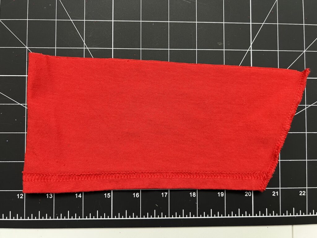 A piece of red t-shirt sleeve fabric with a serged edge lies on a black cutting mat marked with a white grid and centimeter measurements, ready to be transformed into handy drawstring bags.