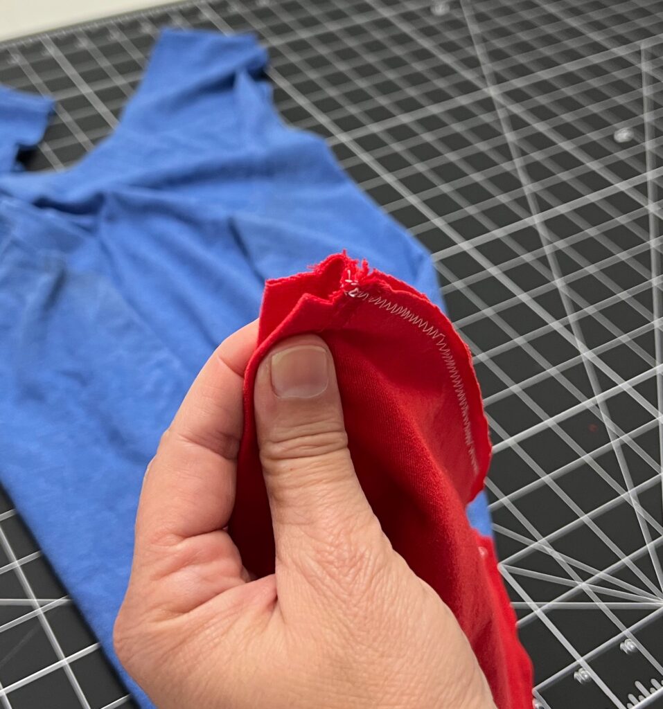 A hand holds the edge of a red seam with zigzag stitches; a blue t-shirt bag and a grid cutting mat are in the background.
