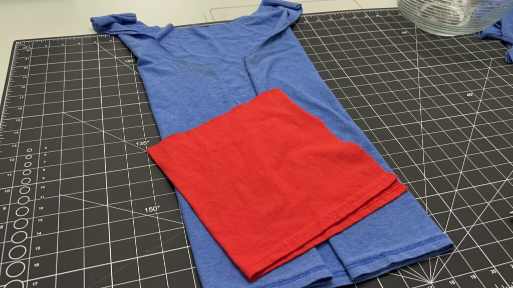 A blue T-shirt with its sleeves inside out is laid on a gridded cutting mat, ready to be transformed into a t-shirt bag, with a folded red fabric piece placed on top.