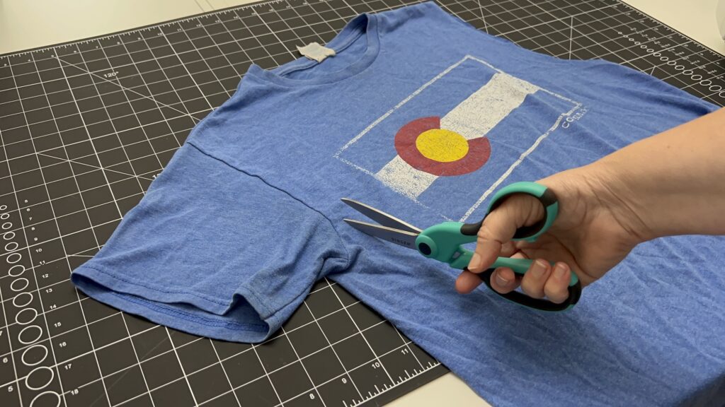 A hand holding scissors prepares to cut the sleeve off a blue Colorado flag T-shirt, laid on a black cutting mat, as the first step in making a DIY t-shirt bag.