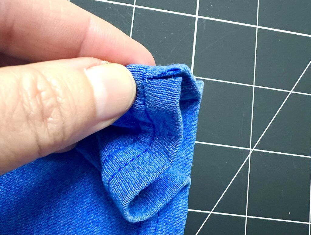 A hand holds the hem of a blue t-shirt sleeve, showing close-up details of the stitching, against a black grid cutting mat—perfect for making drawstring bags.
