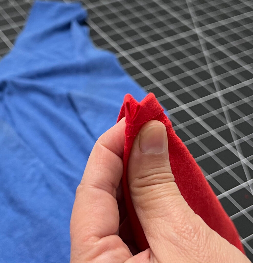 A hand holds the edge of a red fabric, possibly destined to become a t-shirt bag, against a grid-patterned background, with a blue cloth lying flat nearby.