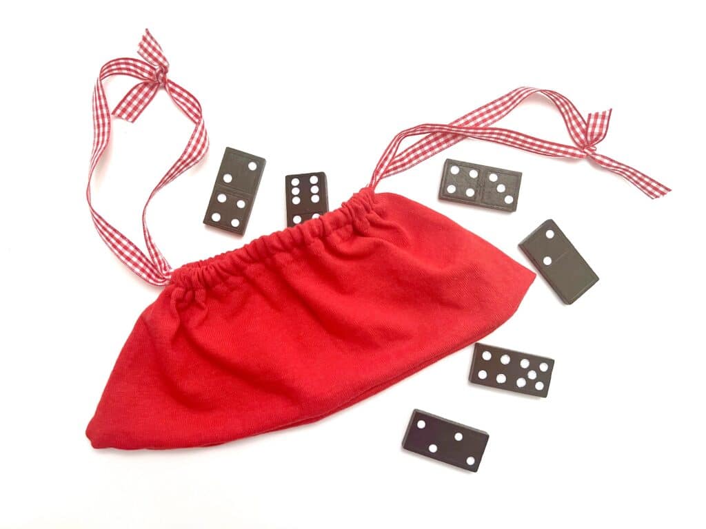 A red t-shirt sleeve drawstring bag with gingham ties is surrounded by several black domino tiles on a white background.