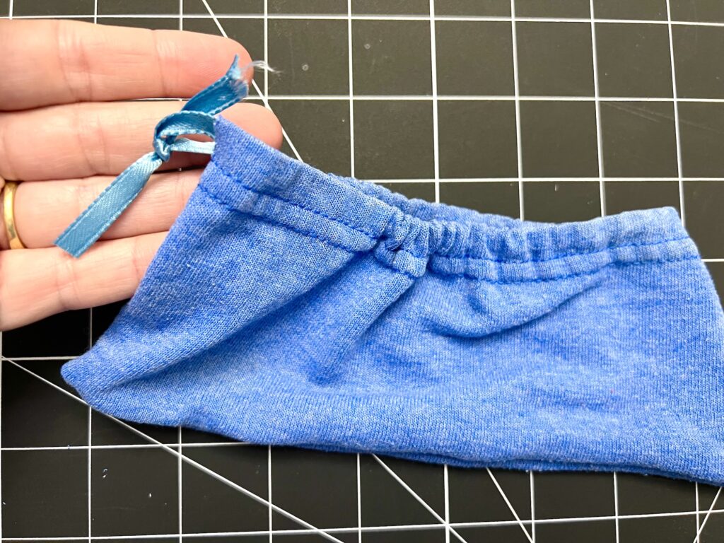 A hand holds a blue t-shirt sleeve drawstring bag with a blue ribbon threaded through the top seam, set against a black grid background.