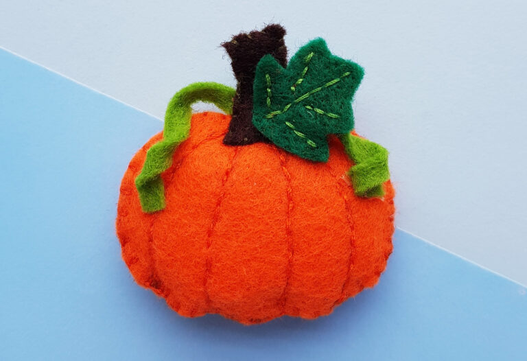 A handmade felt pumpkin with a brown stem, two green vines, and a green leaf displayed against a two toned blue background