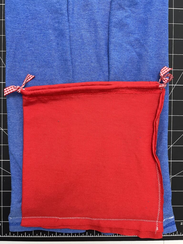 A red rectangular fabric piece with two tied corners, resembling a t-shirt bag in progress, sits atop a larger blue fabric piece on a black grid cutting mat with a visible ruler.
