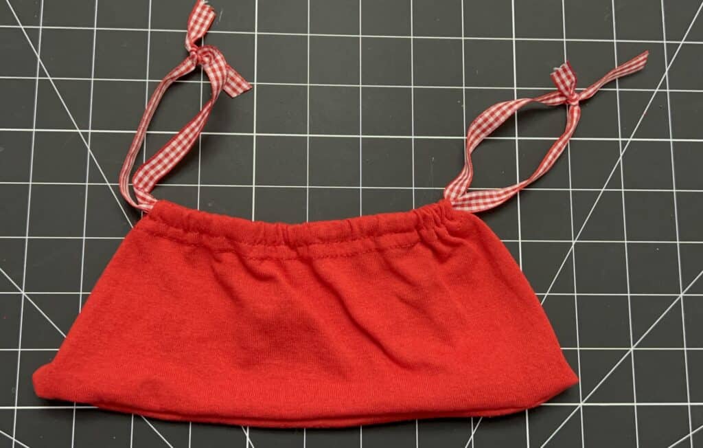 A small red fabric pouch, reminiscent of t-shirt sleeve drawstring bags, with a gathered top and two red gingham ribbon ties, placed on a black grid cutting mat.