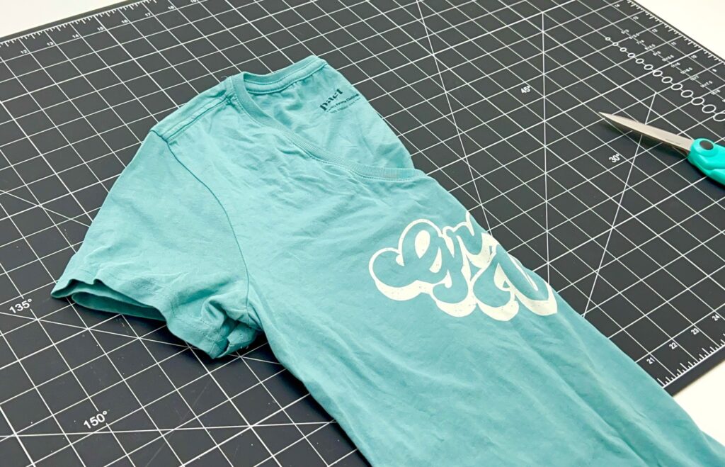 A teal graphic T-shirt is laid on a black grid cutting mat with a pair of blue-handled scissors nearby, ready to be transformed into a stylish t-shirt bag.