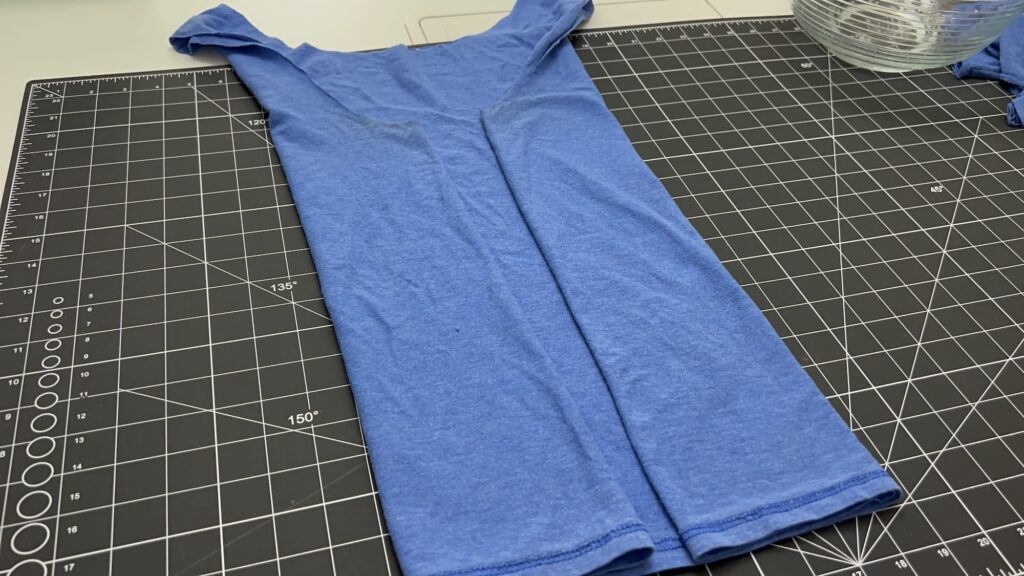 A blue T-shirt, neatly folded lengthwise to prep for making a t-shirt bag, sits on a black cutting mat with white gridlines and measurement markings.