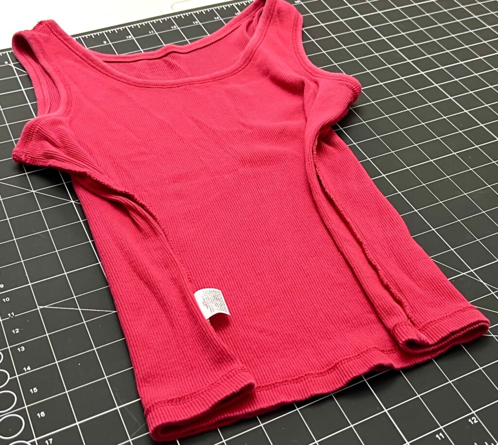 A red ribbed tank top with open side seams is laid flat on a gridded cutting mat, showing an exposed care label—an ideal piece for transforming into a DIY t-shirt bag.