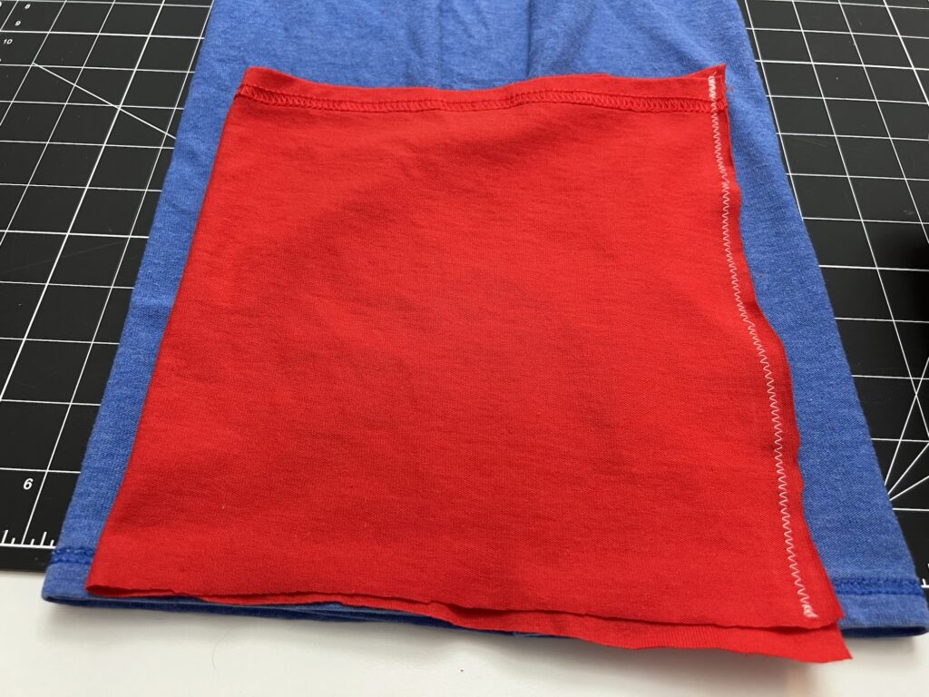 A red fabric rectangle with zigzag stitching on one edge, possibly part of a t-shirt bag project, is placed on top of a blue fabric over a cutting mat with a grid pattern.