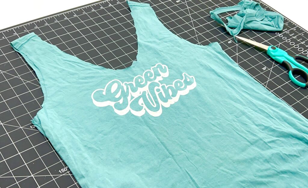A turquoise shirt with "Green Vibes" text is transformed into a t-shirt bag and cut into a tank top on a grid cutting mat, with scissors and fabric scraps beside it.