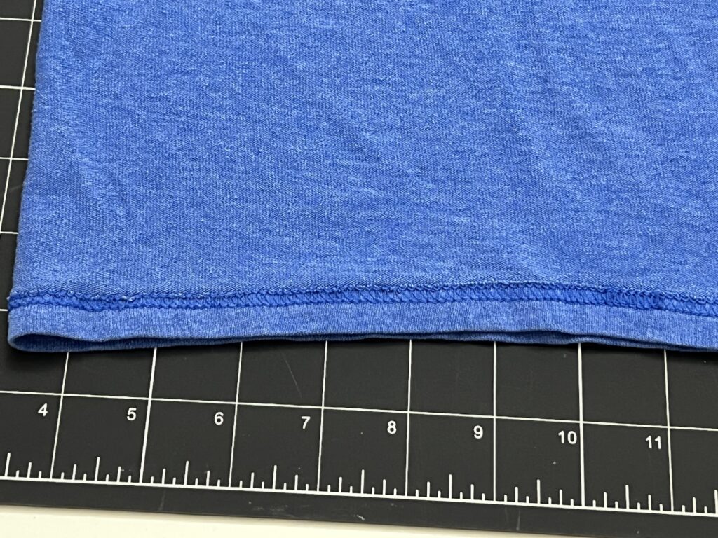 A close-up of the hem of a blue fabric t-shirt bag laid on a black cutting mat with white measurement markings in inches.