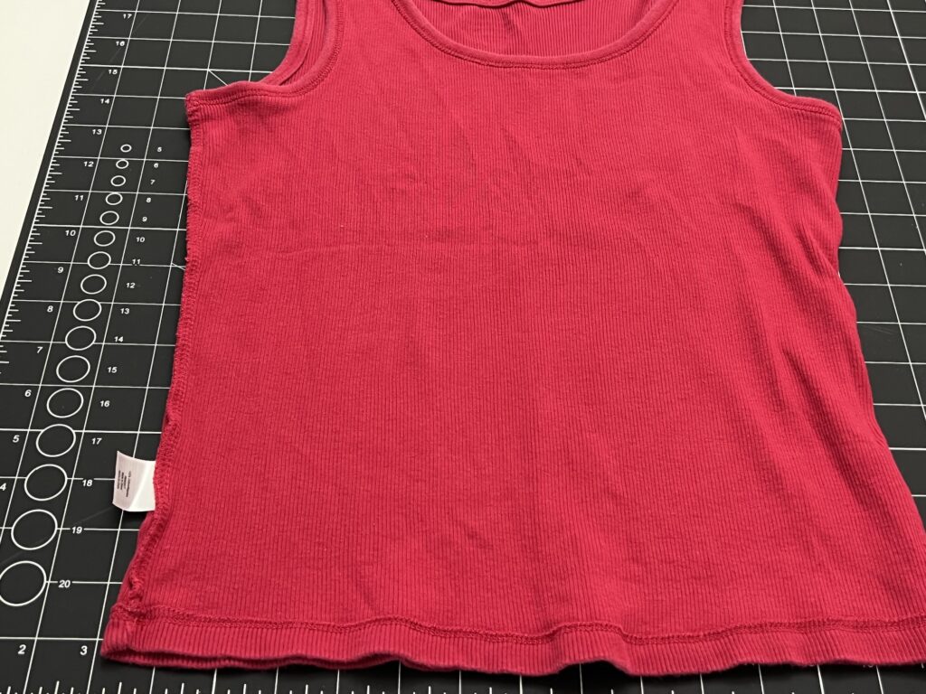 A sleeveless red ribbed tank top, perfect for pairing with a t-shirt bag, is laid flat on a black measurement grid cutting mat, with a white tag visible on the left side seam.