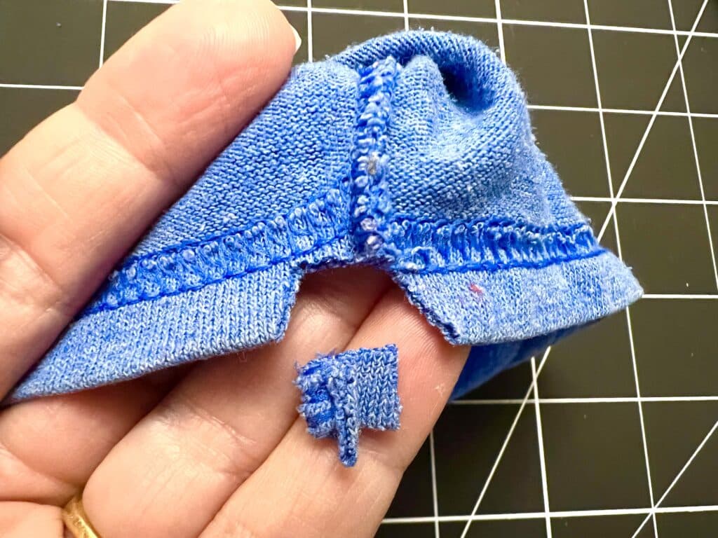 A hand holds a blue knit t-shirt sleeve with a small matching cut-out piece, showing the hem and stitching detail against a black grid background—perfect for crafting drawstring bags.