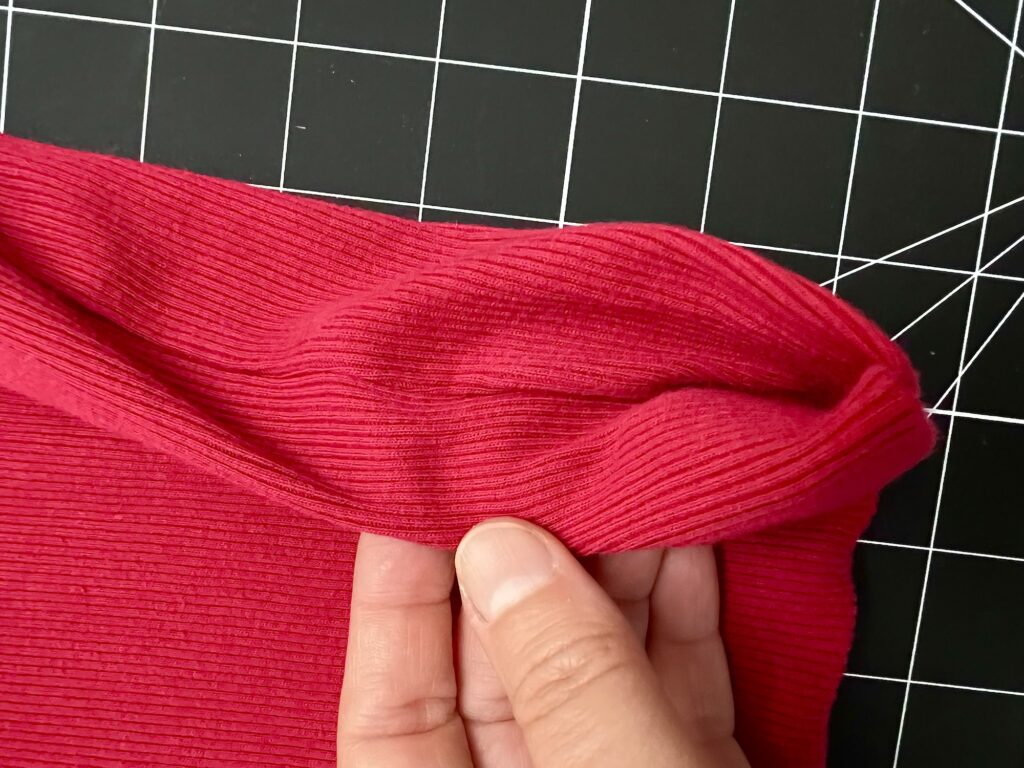 A hand pinches and stretches the edge of ribbed red fabric—soon to become a t-shirt bag—on a black and white grid cutting mat.