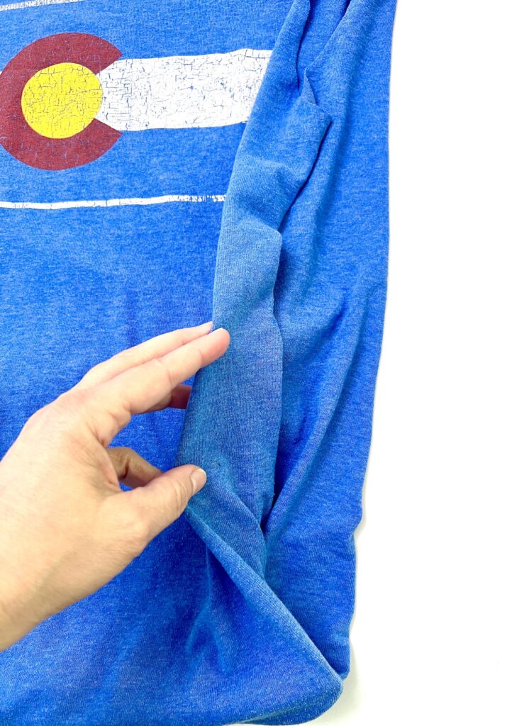 A hand pinches blue fabric with a worn Colorado state flag print, highlighting the texture and stretch of this unique t-shirt bag against a white background.