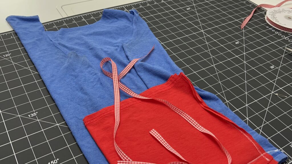 A blue T-shirt and a red fabric piece with checkered ribbon ties, perfect for crafting a t-shirt bag, are laid out on a black grid cutting mat with a roll of ribbon nearby.