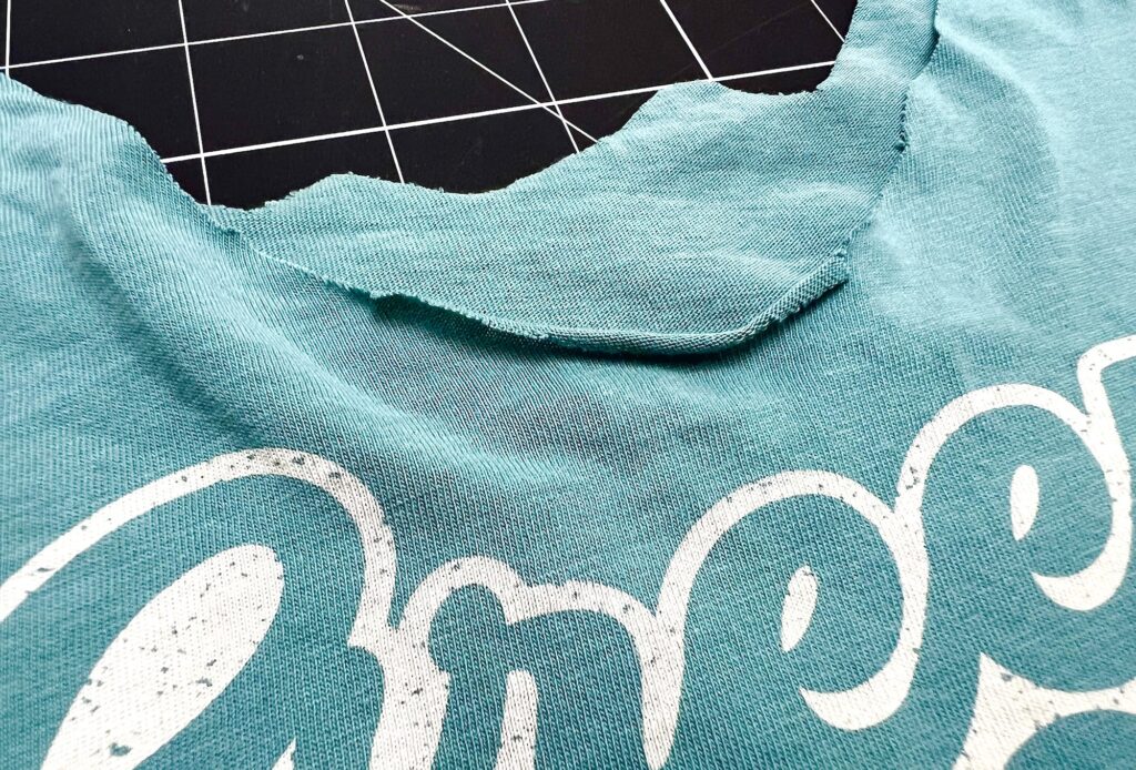 A close-up of a blue-green T-shirt with a roughly cut neckline is displayed on a black grid cutting mat, ready to be transformed into a t-shirt bag.