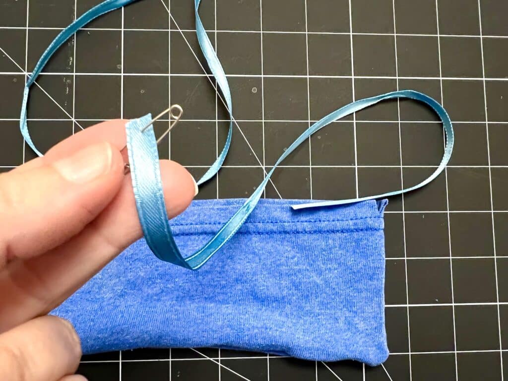 A hand holds a blue ribbon with a safety pin attached, next to a blue fabric piece—perfect for crafting t-shirt sleeve drawstring bags—on a black grid cutting mat.