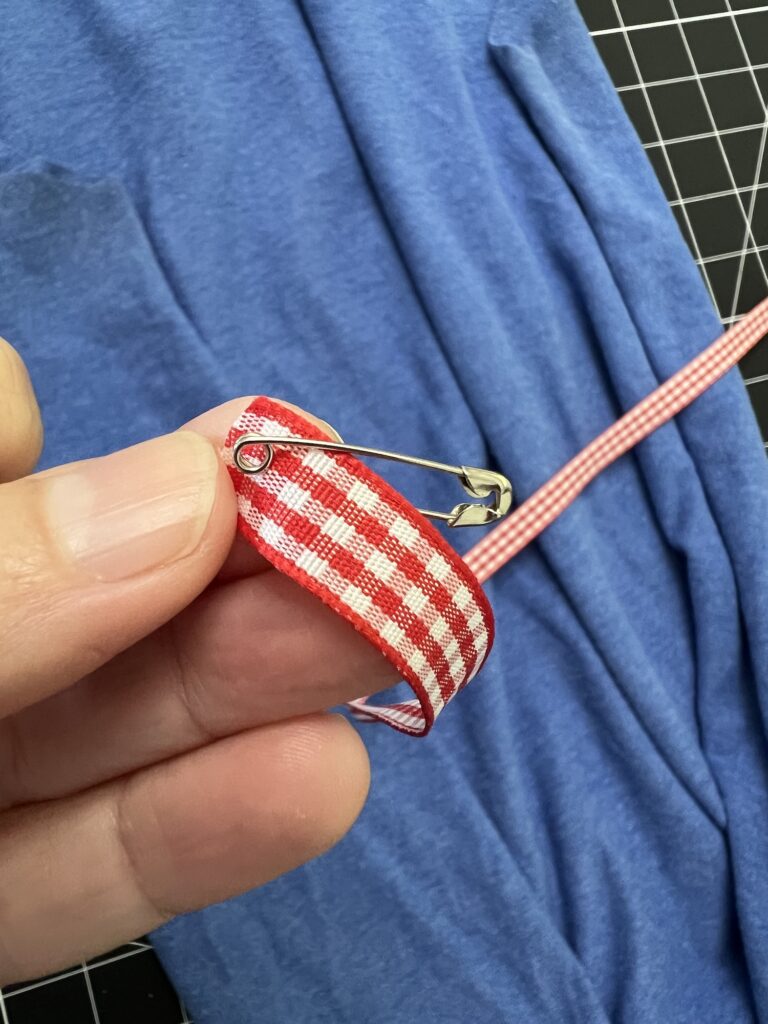 A hand holds a red and white plaid ribbon with a safety pin attached, set against blue fabric—perhaps from a t-shirt bag—and a black grid cutting mat.