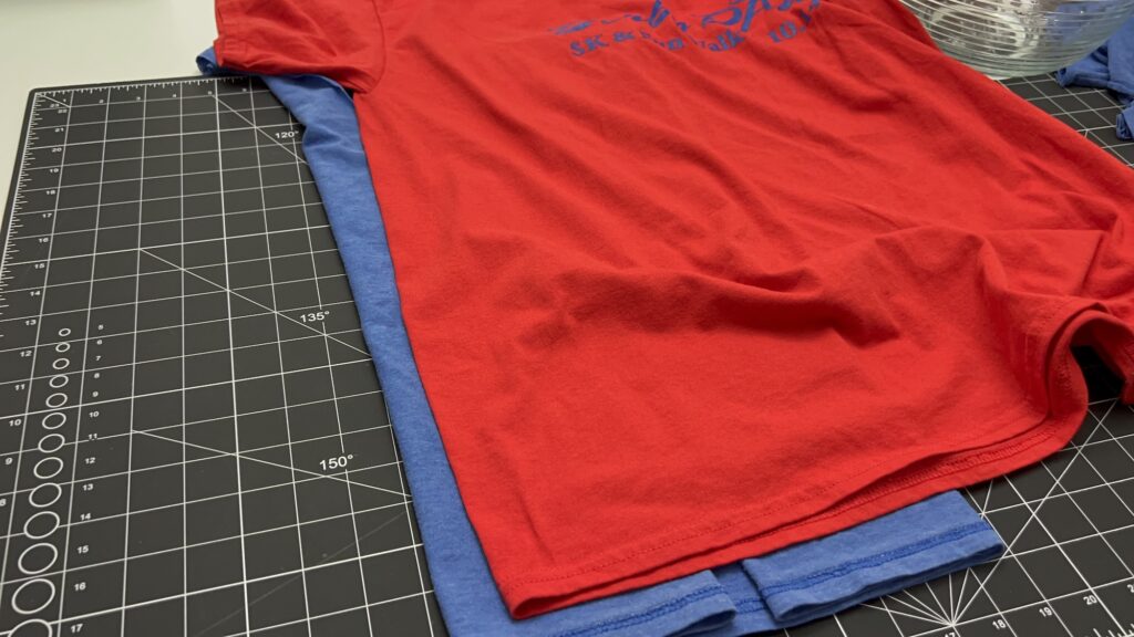 A red t-shirt is stacked on top of a blue t-shirt on a black cutting mat with measurement markings, ready to be upcycled into a t-shirt bag.