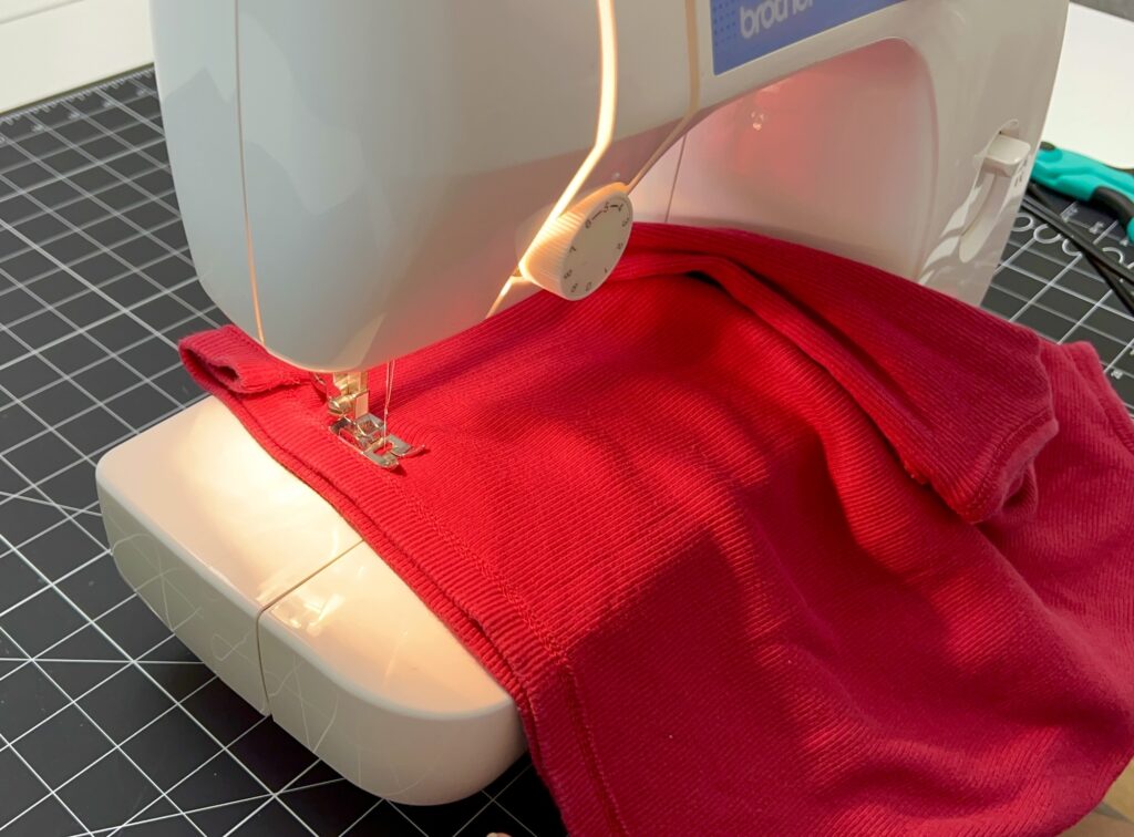 A sewing machine is stitching red fabric on a black grid-patterned cutting mat, possibly creating a t-shirt bag, with scissors visible in the background.