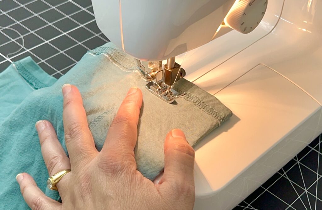 A hand guiding green fabric through a sewing machine, stitching along a seam under bright light—crafting a t-shirt bag with precision and care.