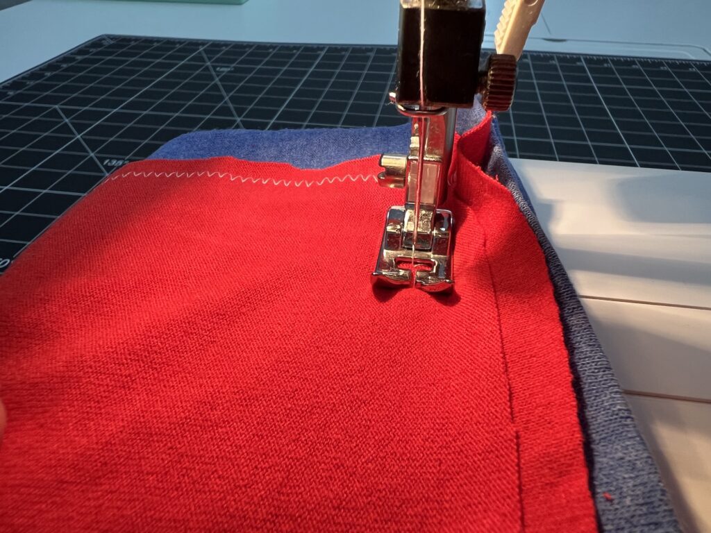 A sewing machine stitches a red t-shirt bag with blue trim on a cutting mat, featuring both zigzag and straight stitches.