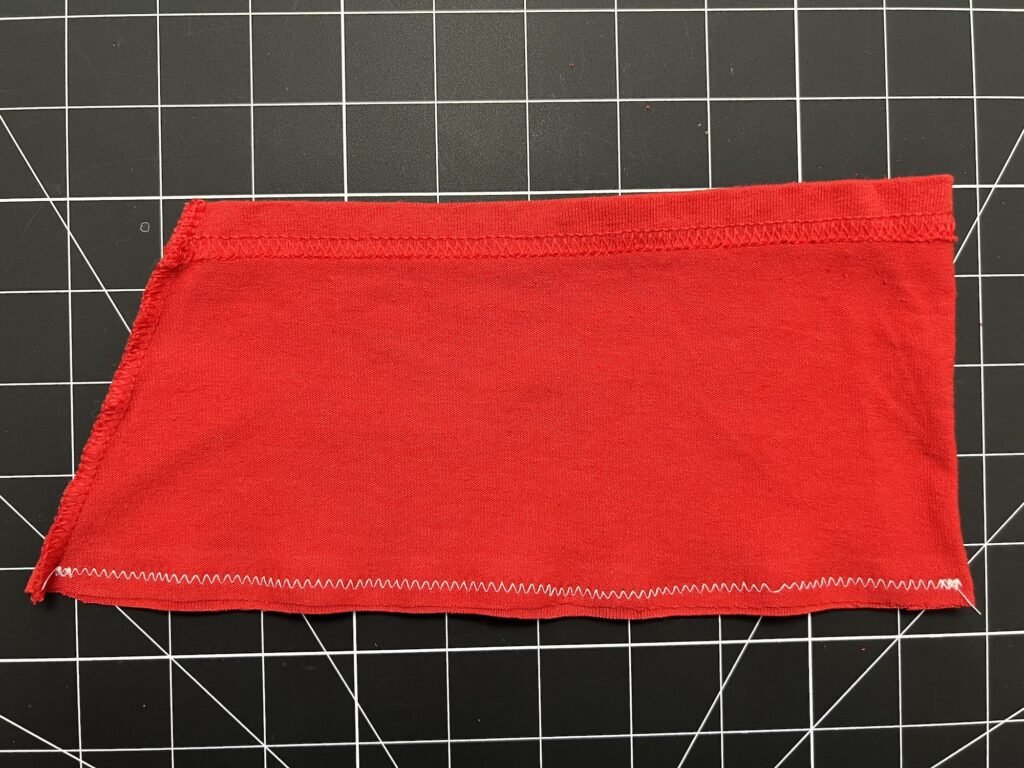 A red piece of fabric, possibly from a t-shirt sleeve, with zigzag stitching on one edge and straight stitching on top, placed on a black and white grid cutting mat—ideal for crafting drawstring bags.